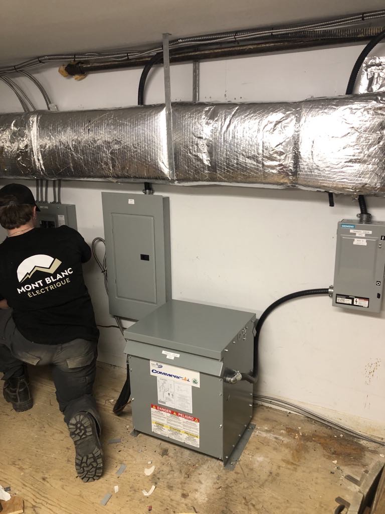 Électricien portant un casque de sécurité et des lunettes de protection, utilisant un tournevis pour travailler sur un tableau électrique avec des câbles visibles dans un environnement industriel.