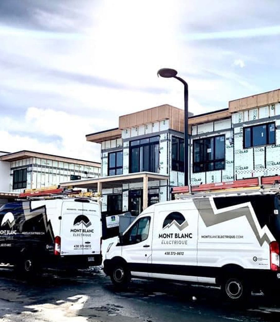 Deux camions de la compagnie Mont Blanc Électrique stationnés devant un bâtiment résidentiel en construction, sous un ciel partiellement nuageux.